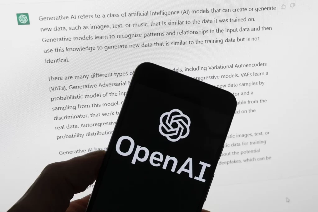 OpenAI 的标志出现在一部手机的屏幕上，该手机放在一台显示 ChatGPT 输出的电脑屏幕前。