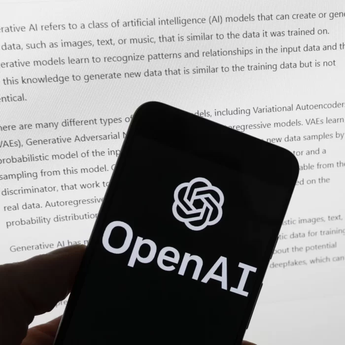 OpenAI 的标志出现在一部手机的屏幕上，该手机放在一台显示 ChatGPT 输出的电脑屏幕前。